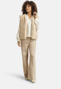 Vrouw staand, gekleed in een beige faux fur-vest, witte blouse met lange mouwen en borduurwerk, beige wijde broekspijpen en beige puntige schoenen.