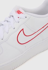 Hvide lædersneakers med en struktureret finish, der har et rødt syet Nike Swoosh-logo og hvide snørebånd, hvilket fremhæver et lavt design.