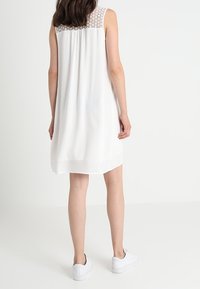 Biała sukienka bez rękawów z teksturowanym koronkowym tyłem. Luźny krój, do kolan, zwiewny materiał, zestawiona z białymi trampkami. Prosty design.