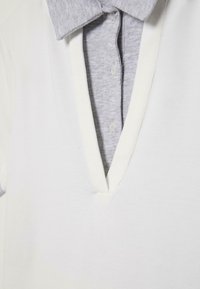 Haut blanc à col en V superposé sur une chemise gris clair à col avec boutons, montrant le col ouvert et les deux premiers boutons de la chemise.