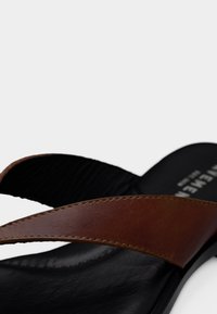 Καφέ δερμάτινα σανδάλια με μαύρη υφασμάτινη σόλα. Οι σταυρωτοί ιμάντες και η λεπτομέρεια με ραφές αναδεικνύουν τον μινιμαλιστικό σχεδιασμό.