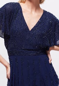 Vestido azul marino con adornos de cuentas en patrones ondulados. Presenta un escote en V y mangas fluidas, cintura entallada y textura en capas.