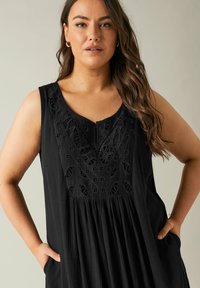 Vestido negro sin mangas de tela texturizada, con intrincados bordados en el escote y un diseño de falda fluida y fruncida con bolsillos.
