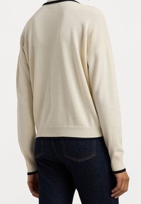 Svetle krémový svetr s volným střihem, žebrovanými manžetami a lemem, a tmavým kontrastním lemováním kolem výstřihu. Nosený přes tmavé džíny.