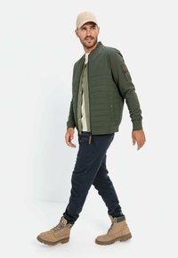 Groene bomberjack met gewatteerde details, gedragen over een lichtgekleurde shirt, in combinatie met donkere broek en tan laarzen. Beige pet complementeert de outfit.