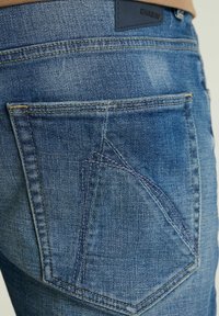 Kék farmernadrág, amely egyedi hátsó zsebdizájnnal rendelkezik, látható varrási részletekkel és a derékpánt felett elhelyezett márkajelzéssel.