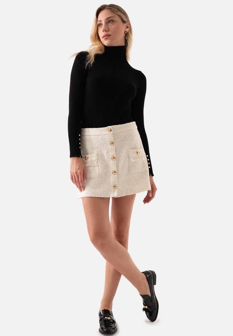 Zwarte getailleerde coltrui, witte bouclé minirok met gouden knoopsluiting en klepzakken, gecombineerd met zwarte loafers. Soepele textuur.