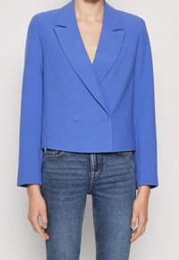 Blazer bleu cropped à manches longues, boutons croisés, col cranté ; texture lisse, associé à un jean foncé.