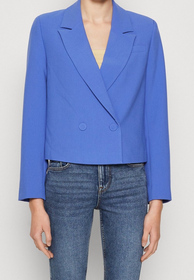 Blazer bleu cropped à manches longues, boutons croisés, col cranté ; texture lisse, associé à un jean foncé.