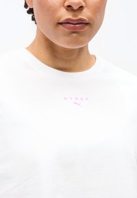 Κοντινό πλάνο ενός ατόμου που φοράει λευκό πουκάμισο με το λογότυπο "HYROX" και το λογότυπο της Puma εκτυπωμένα σε ανοιχτό ροζ στο στήθος.