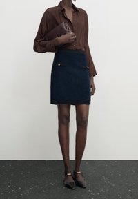 Brun knappad skjorta med uppvikta ärmar, ihop med en strukturerad marinblå minikjol med svarta detaljer och gyllene knappar. Svarta spetsiga skor.