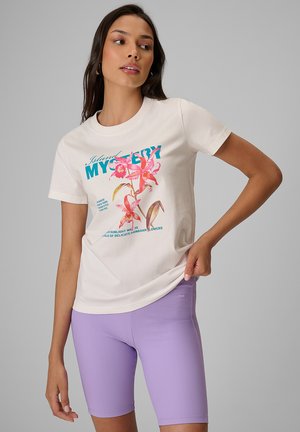 Kobieta w kremowym t-shircie z różowymi kwiatami i niebieskim napisem, zestawionym z lawendowymi, prążkowanymi kolarkami z wysokim stanem, stoi na neutralnym tle.
