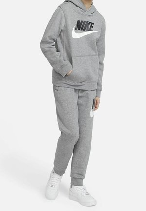 Kind draagt een grijze Nike-hoodie en trainingsbroek met witte Nike-sneakers, staat met één hand in de zak tegen een witte achtergrond.