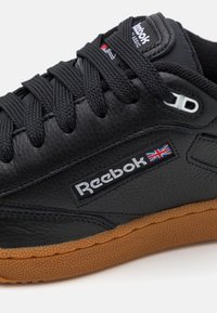 Baskets en cuir noir avec finition texturée, arborant un logo Reebok blanc, une semelle en caoutchouc gomme et une fermeture à lacets noire.