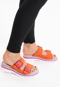 Sandali in suede arancione con due grandi fibbie quadrate rosa, dotati di una suola bianca con un accento viola. Indossati con leggings neri.