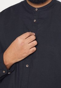Camicia nera con colletto alla coreana, maniche lunghe e chiusura con bottoni. Il tessuto ha una superficie liscia con dettagli di cucitura visibili.