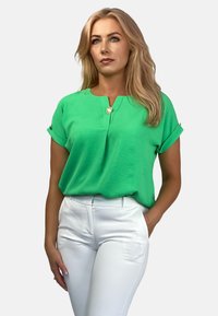 Zielona bluzka z krótkim rękawem w serek z ozdobnym guzikiem przy kołnierzy, zestawiona z dopasowanymi białymi spodniami. Gładka faktura.