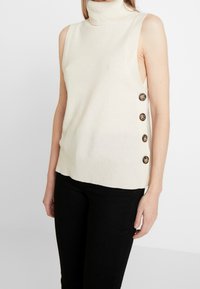 Ärmlös polotröja i off-white, med sidoknappar, ribbad fåll och en mjuk textur. Kombinerad med svarta byxor.