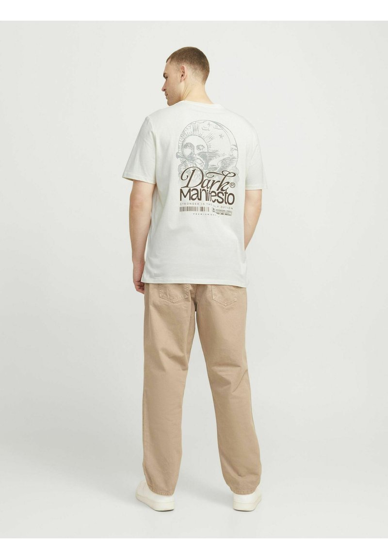 Hvid bomulds t-shirt med et grafisk print på ryggen med teksten "Dark Manifesto" og illustrationer. Sammen med beige afslappede bukser.