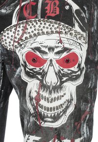 Schwarze Denim-Hose mit einem großen Totenkopf-Design in roten Akzenten und metallischen Nieten. Zerrissene Struktur mit Farbspritzern.