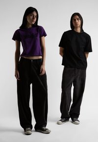 Una maglietta viola corta con testo grafico nero abbinata a pantaloni larghi di velluto a coste neri e scarpe da ginnastica nere. Colori solidi, vestibilità comoda.