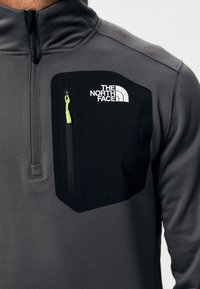 Pull-over en polaire gris avec poche poitrine noire, doté d'une fermeture éclair et d'une tirette de fermeture éclair verte. Le logo The North Face est affiché de manière proéminente.