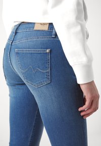 Jeans en denim bleu ajustés présentant une finition texturée, des poches arrière visibles avec des coutures en forme de vagues, et une étiquette de ceinture en cuir beige.