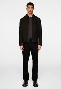 Schwarze Reißverschlussjacke mit strukturiertem Kragen, getragen über einem dunklen Hemd, kombiniert mit geraden schwarzen Hosen und schwarzen Schuhen. Glatte Stoffstruktur.