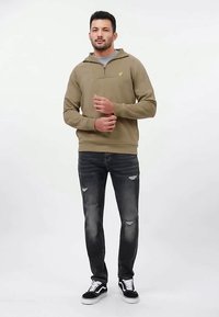 Hanorac kaki cu fermoar și finisaj texturat, purtat peste un top gri, asortat cu blugi întunecați rupți �și adidași negri. Are un detaliu cu logo.