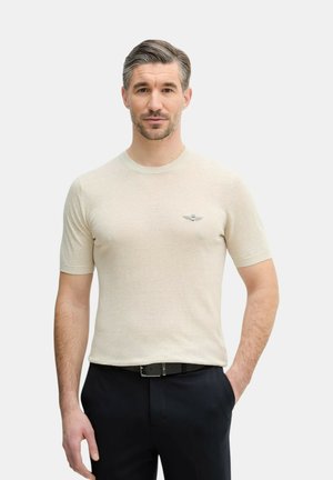 Uomo con capelli corti sale e pepe che indossa un maglione beige a maniche corte e pantaloni neri, in piedi con una mano in tasca.