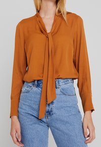 Brun langærmet bluse med bånd ved halsen; glat stof; parret med højtaljede blå denimjeans.