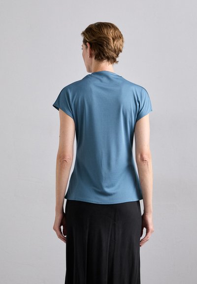Kortermet, blå T-skjorte laget av mykt stoff, tettsittende fasong, med en glatt tekstur. Båret sammen med et langt svart skjørt, minimalistisk design.