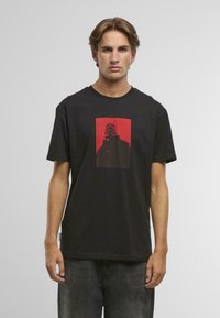 Mladý muž s krátkymi svetlohnedými vlasmi, ktorý má na sebe čierne tričko s červeným obdĺžnikovým grafickým motívom osoby v tmavom oblečení.