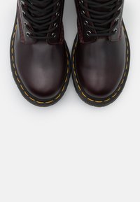 Botins de couro burgundy até ao tornozelo com atacadores pretos, bico redondo e costura amarela na sola de borracha preta. Detalhe de superfície texturizada.