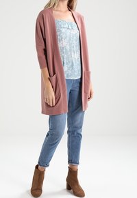 Nadměrný, žebrovaný svetřík světle růžové barvy s dvěma předními kapsami, nošený přes modré květinové tílko, kombinovaný se světle modrými džíny a hnědými kotníkovými botami.