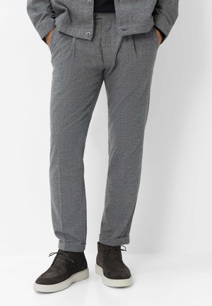 Man draagt grijze op maat gemaakte broek en donkerbruine suède enkellaarsjes met witte zolen, staand met handen in de zakken.