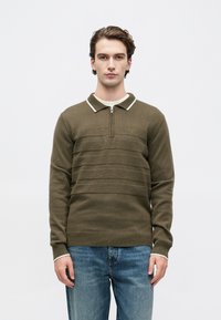 Jovem vestido com um suéter verde-oliva com fecho e colarinho branco e calças de ganga azuis, de pé contra um fundo branco liso.