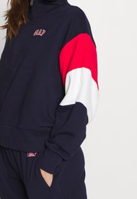 Pullover in pile blu navy con colletto alto, chiusura con zip e accenti diagonali colorati sulle maniche in rosso e bianco. Logo ricamato sul petto.