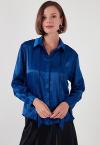 Long-sleeve shirt in shiny blue satin with a collar, button front, and tying detail at the waist; smooth texture and relaxed fit.