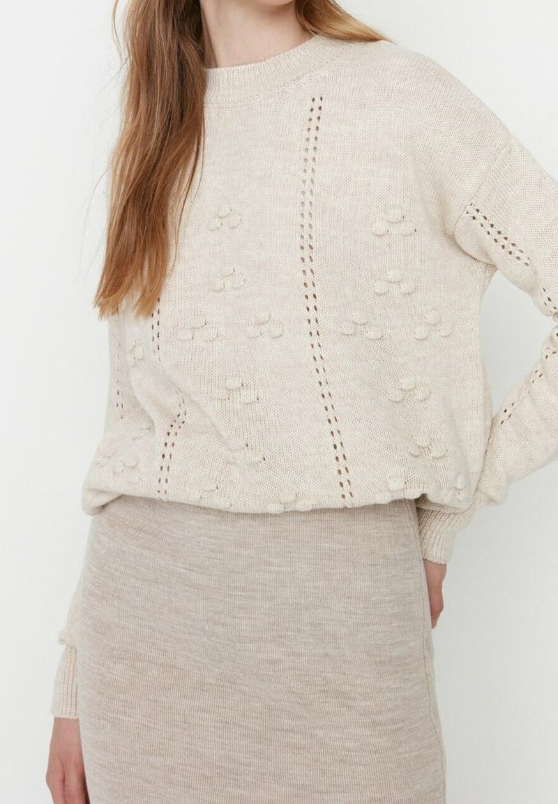 Femme portant un pull en maille beige avec des motifs en relief et une jupe assortie en maille côtelée, posant avec une main sur la hanche.