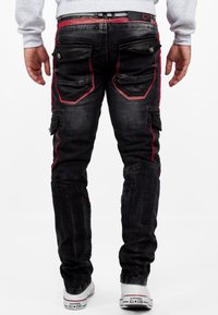 Jeans noirs avec des poches de style cargo, des accents rouges le long des coutures et des détails en simili cuir. Portés avec des baskets blanches.