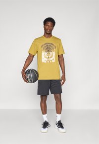 Kortermet t-skjorte i sennepsgul med grafisk trykk, kombinert med svarte shorts og svart-hvite athletic sko, holdende en basketball.
