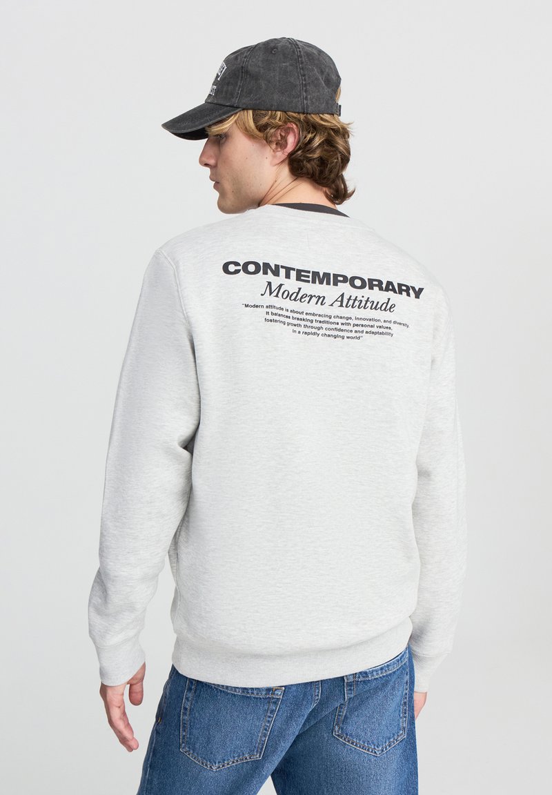 Felpa grigia con maniche lunghe e polsini a costine, con testo nero sul retro. Abbinata a jeans blu. Cappellino scuro regolabile indossato al contrario.