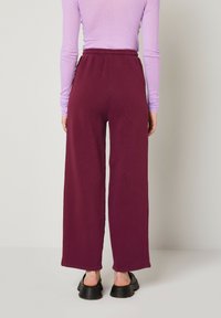 Tumši sarkani plašas bikses ar elastīgu jostu, izgatavotas no mīksta auduma, apvienotas ar gariem piedurknēm purpursarkanā augšpusē un melnu apaviem.