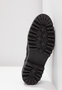 Botte noire avec semelle en caoutchouc épaisse à sculptures profondes, présentant des crampons profonds et des motifs en forme de croix, sur une surface d'angle blanche.
