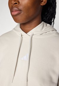 Beige hoodie with a drawstring, featuring a white Adidas logo on the chest. Smooth texture with a relaxed fit.