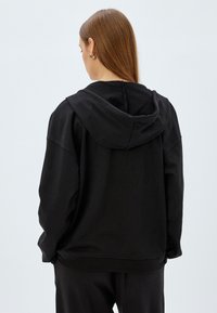 Sudadera negra con un corte amplio, que presenta una gran capucha, puños y dobladillo acanalados, y una textura de tela suave, vista desde la parte posterior.