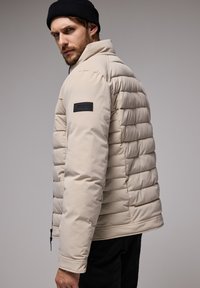 Beige Puffjacke mit horizontalem Steppmuster, Reißverschluss und schwarzem Etikett am linken Ärmel. Glatte Textur, leicht taillierter Schnitt.