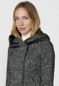 Čierno-biele škvrnité sako s veľkou kapucňou, textúrovaný materiál a predný zips. Obsahuje vysoký golier a priliehajúci strih.