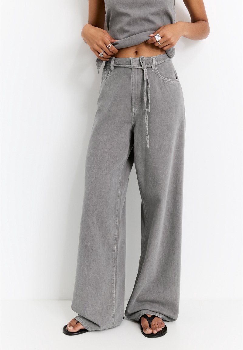 Personne portant un pantalon large gris ample avec une taille à cordon et des sandales noires à bout ouvert, soulevant un haut gris pour révéler la taille.